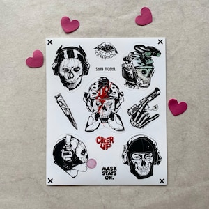Puede incluir: Una hoja de pegatinas con ilustraciones en blanco y negro de personajes con calaveras, equipo t&aacute;ctico y armas. El texto incluye "STAY FROSTY", "GHOST", "CHEER UP" y "MASK STAYS ON". Los dise&ntilde;os est&aacute;n detallados con l&iacute;neas de tinta de alto contraste.