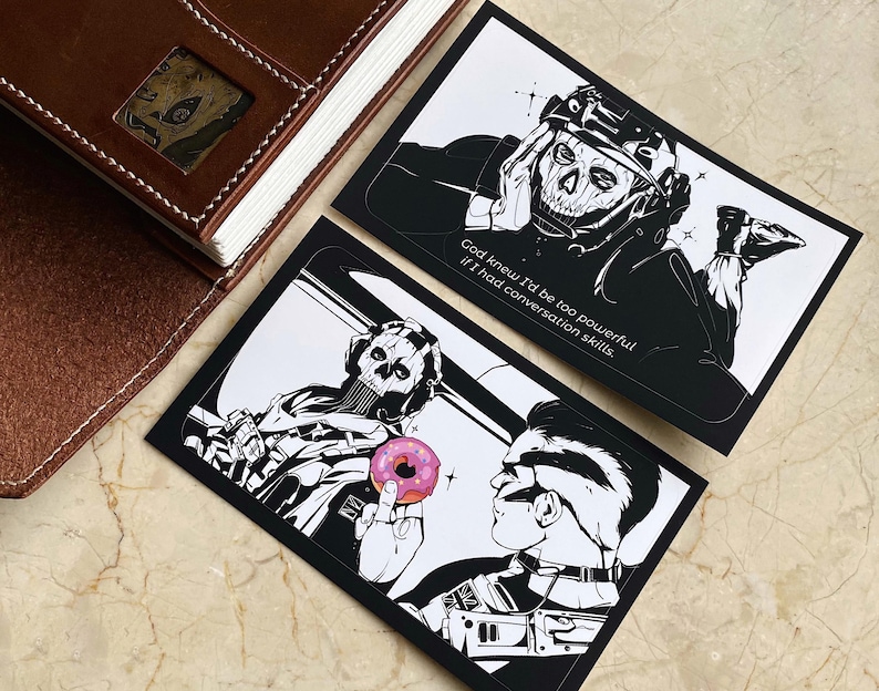 K&ouml;nnte beinhalten: Zwei rechteckige Kunstdrucke mit schwarz-wei&szlig;en Illustrationen einer Figur mit Totenkopfmaske. Ein Druck zeigt die Figur mit dem Text, God knew I'd be too powerful if I had conversation skills. Der andere zeigt einen rosa Donut.