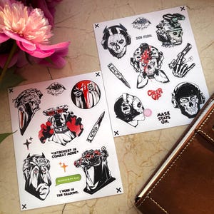 May include: Two sticker sheets featuring black and white illustrations of skulls, helmets, and weapons. One sheet includes text that says "INTROVERT IN COMBAT MODE", "SILENCE IS MY ALLY", and "I WORK IN THE SHADOWS." The other sheet says "CHEER UP" and "MASK STATS ON."