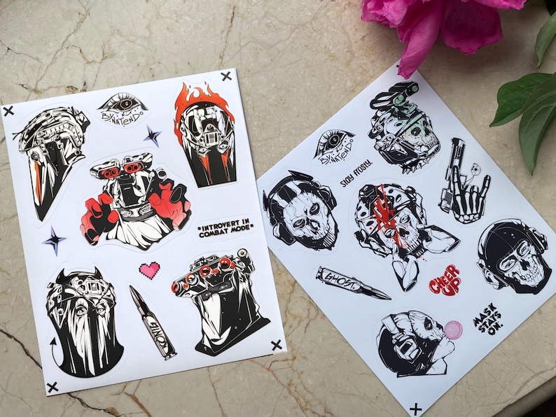 Puede incluir: Dos hojas de pegatinas blancas con ilustraciones gr&aacute;ficas en negro, blanco y rojo de equipo t&aacute;ctico, calaveras y personajes militares. El texto incluye "INTROVERT IN COMBAT MODE", "STAY FROSTY", "GHOST", "CHEER UP" y "MASK STAYS ON".