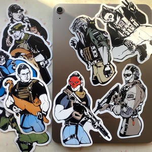 May include: A collection of die-cut vinyl stickers featuring stylised illustrations of tactical characters in military gear, some accompanied by cats. The stickers display various figures in combat attire, including one with a red skull mask and rifle.