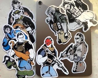 Puede incluir: Una colección de pegatinas de vinilo troqueladas con ilustraciones estilizadas de personajes tácticos con equipo militar, algunos acompañados de gatos. Las pegatinas muestran varias figuras con ropa de combate, incluida una con una máscara de calavera roja.