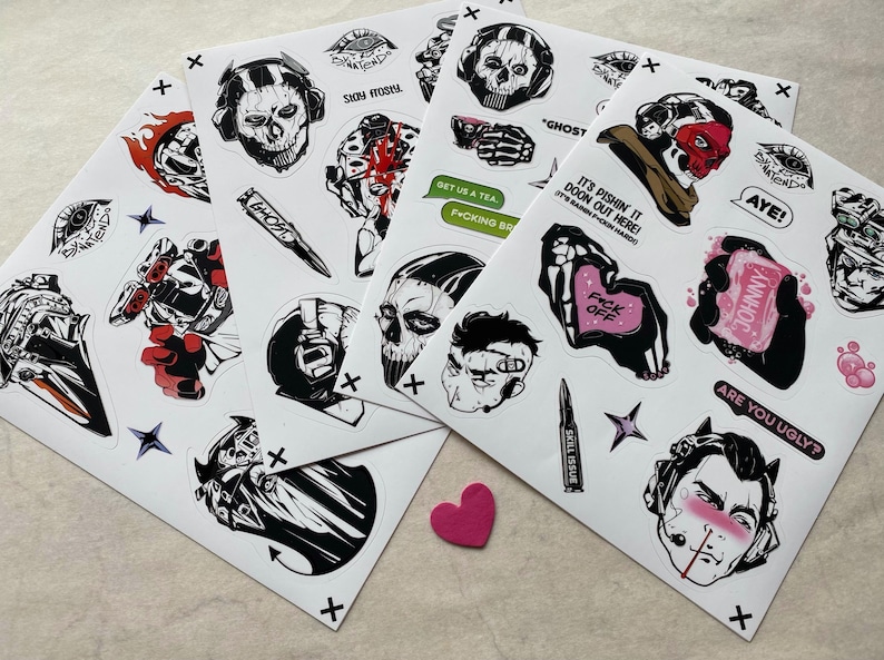 Puede incluir: Una colecci&oacute;n de hojas de pegatinas con diversas ilustraciones gr&aacute;ficas, incluyendo personajes con m&aacute;scaras de calavera, motivos militares y elementos de texto como "STAY FROSTY", "GHOST", "GET US A TEA", "F*CK OFF", "JOHNNY" y "ARE YOU UGLY?" en negro, blanco y rojo.