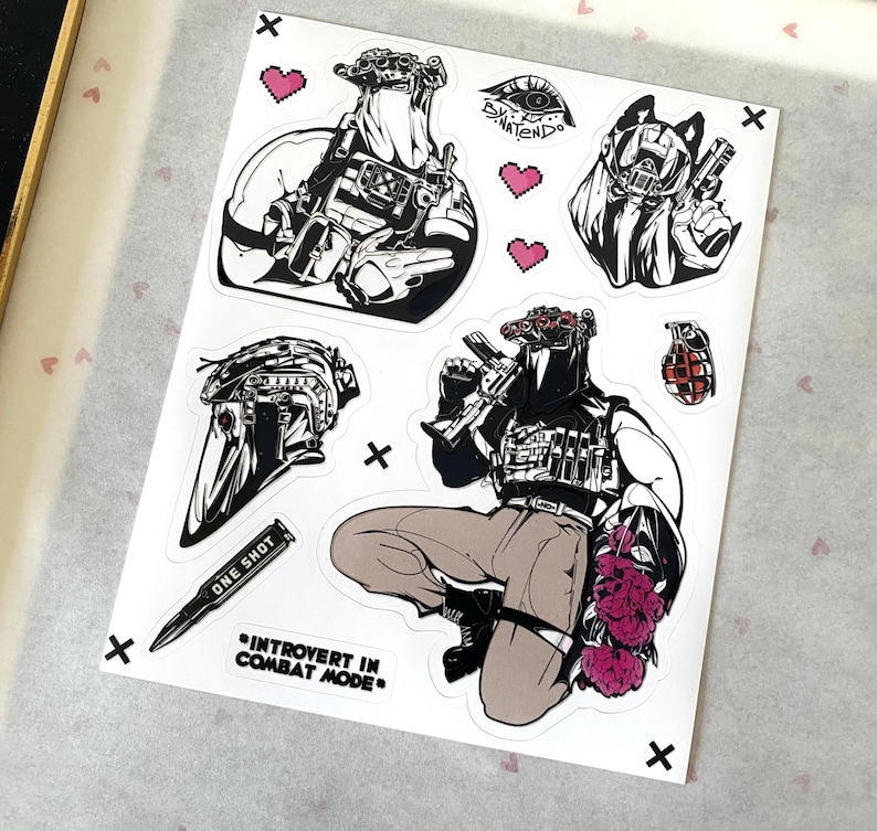 Puede incluir: Una hoja de pegatinas con ilustraciones t&aacute;cticas en blanco y negro, que incluyen figuras con equipo de combate, un casco, una granada, una bala con la etiqueta ONE SHOT y el texto INTROVERT IN COMBAT MODE, con peque&ntilde;os corazones rosas pixelados.