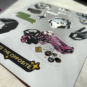 Puede incluir: Una hoja de pegatinas con varios dise&ntilde;os, incluyendo una pistola rosa con peque&ntilde;os dijes de personajes, una pegatina negra con el texto QUITE THE OPPOSITE, una calavera, un signo de paz y otros elementos gr&aacute;ficos con acabado brillante.