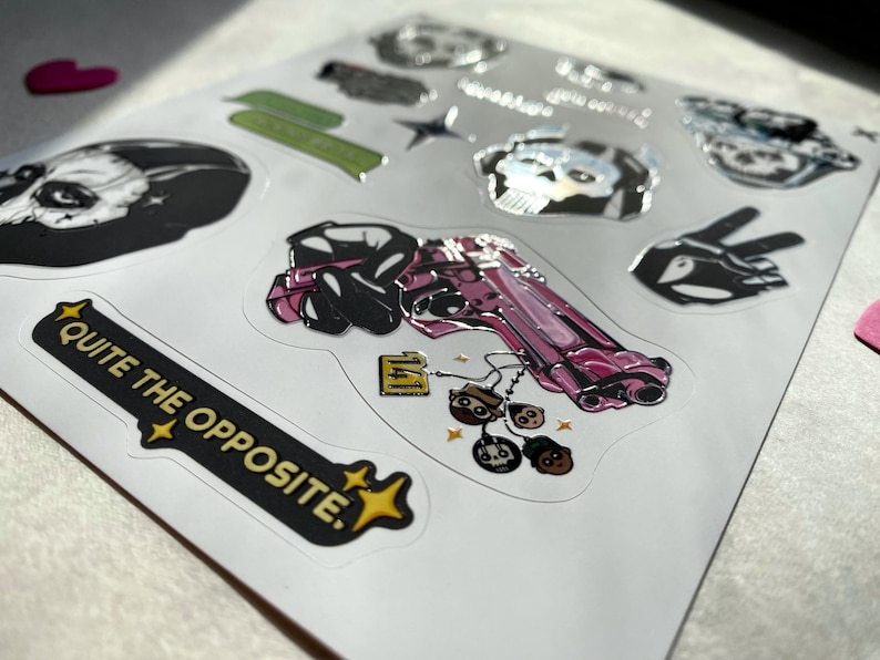 Puede incluir: Una hoja de pegatinas con varios dise&ntilde;os, incluyendo una pistola rosa con peque&ntilde;os dijes de personajes, una pegatina negra con el texto QUITE THE OPPOSITE, una calavera, un signo de paz y otros elementos gr&aacute;ficos con acabado brillante.