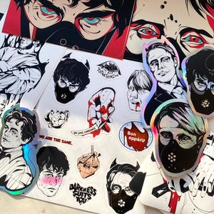 Puede incluir: Una colección de pegatinas e impresiones con ilustraciones estilizadas. La obra de arte incluye retratos, diseños gráficos y texto, con una mezcla de elementos negros, blancos, rojos y holográficos. Algunas pegatinas tienen el texto "Bon appétit" y "Darkness Suits You".