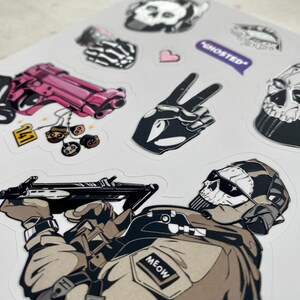 Puede incluir: Una hoja de pegatinas con varias ilustraciones, incluyendo una pistola rosa con una etiqueta 141, una mano esquel&eacute;tica haciendo el signo de la paz, una m&aacute;scara de calavera, un globo de texto morado con el texto GHOSTED y un soldado enmascarado.