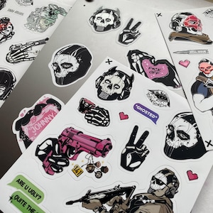 Puede incluir: Varias hojas de pegatinas blancas con diseños de calaveras, armas y frases como "F*ck Off" y "Ghosted". Las pegatinas son en negro, rosa y blanco, algunas incluyen corazones y el texto "Johnny."