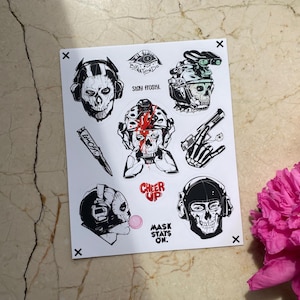 Puede incluir: Una hoja de pegatinas blanca con ilustraciones en blanco y negro de personajes con m&aacute;scaras de calavera y equipo t&aacute;ctico. Los dise&ntilde;os incluyen una bala con la etiqueta GHOST, textos como STAY FROSTY, CHEER UP, MASK STAYS ON y una burbuja rosa.