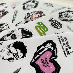 Puede incluir: Una colecci&oacute;n de pegatinas de vinilo troqueladas sobre hojas blancas con ilustraciones gr&aacute;ficas en blanco y negro de calaveras, equipo t&aacute;ctico y personajes. El texto incluye "GET US A TEA", "F*CKING BR" y "IT'S PISHIN' IT DOON OUT HERE!"