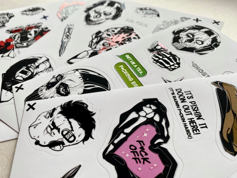 Puede incluir: Una colecci&oacute;n de pegatinas de vinilo troqueladas sobre hojas blancas con ilustraciones gr&aacute;ficas en blanco y negro de calaveras, equipo t&aacute;ctico y personajes. El texto incluye "GET US A TEA", "F*CKING BR" y "IT'S PISHIN' IT DOON OUT HERE!"