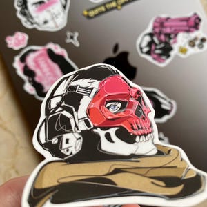 Puede incluir: Una pegatina de vinilo troquelada que muestra un personaje estilizado con una m&aacute;scara de calavera roja y auriculares t&aacute;cticos negros. El personaje lleva una bufanda de color canela alrededor del cuello. El dise&ntilde;o usa contornos negros y rojo.
