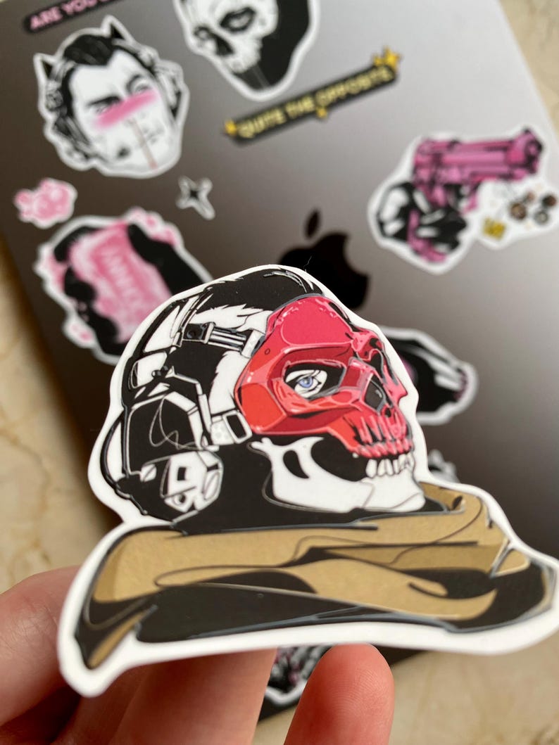 Puede incluir: Una pegatina de vinilo troquelada que muestra un personaje estilizado con una m&aacute;scara de calavera roja y auriculares t&aacute;cticos negros. El personaje lleva una bufanda de color canela alrededor del cuello. El dise&ntilde;o usa contornos negros y rojo.