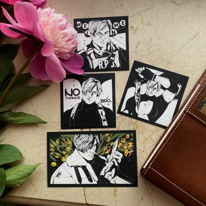 May include: Four black and white stickers featuring illustrations of a character with text. The stickers are arranged on a surface with a brown leather notebook and a pink peony flower. The text on the stickers includes "WELCOME", "R.P.", "NO THANKS", and "BRO."