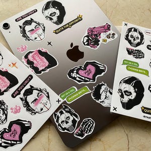 Puede incluir: Un conjunto de pegatinas con varios diseños, incluyendo una calavera, una pistola rosa y un corazón con la frase "F*ck Off". Otras pegatinas muestran texto como "Are You Ugly?" y "Johnny".