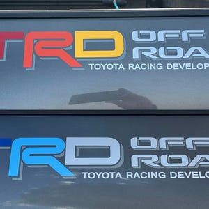 May include: Two close-up images of a vehicle's rear window with a "TRD Off Road" decal. The top image features a red, yellow, and white decal. The bottom image features a blue, white, and gray decal. Both decals include the text "Toyota Racing Development."