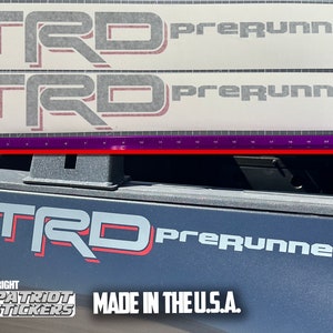May include: Two white vinyl decals with black and red lettering that reads "TRD PreRunner". The decals are stacked on top of each other. Below the decals is a black truck tailgate with a similar decal applied to it. The decal on the tailgate has a red outline. The tailgate also has a sticker that reads "Copyright Patriot Stickers Made in the U.S.A."