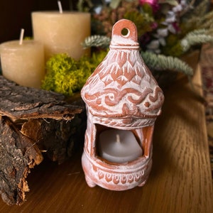 Peut inclure: Un photophore en terre cuite avec une finition blanchie. Le support décoratif en forme de lanterne présente une façade ouverte pour afficher une bougie allumée. Le design comprend des détails sculptés et une petite boucle en haut pour suspendre.