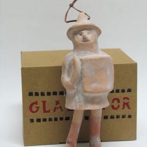 May include: A terracotta-colored figurine of a person wearing a hat and coat, holding a book. The figurine has a leather string for hanging. Behind it is a cardboard box with the word "GLADIATOR" printed in red.