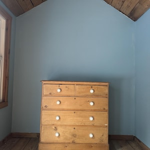 May include: A light brown wooden dresser with five drawers and white knobs. The dresser is in a room with blue walls and a wooden ceiling and floor. The room has a window on the left side.