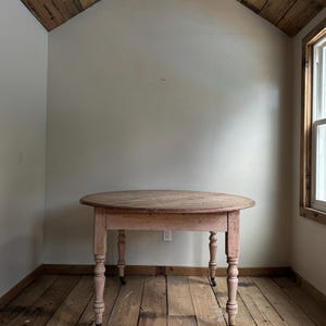 May include: A round, antique wooden table with a light brown finish. The table has four turned legs with small wheels and a distressed appearance. The table is set in a room with wooden floors and a light-colored wall.