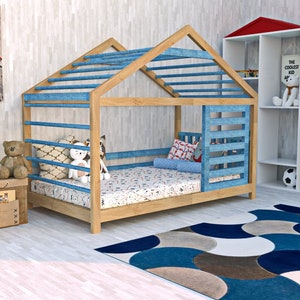 Puede incluir: Una cama de madera con forma de casa con una cubierta de tela azul y un colchón con estampado blanco y azul. La cama está en una habitación infantil con una caja de juguetes de madera, animales de peluche y estantes blancos con libros y juguetes.