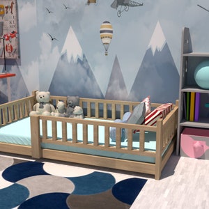 May include: A light wood toddler bed with a blue and white patterned rug, a grey bookshelf with two pink storage bins, and a blue and white hot air balloon on a wall with a mountain scene.