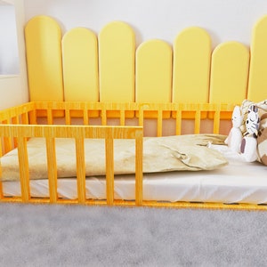 May include: A wooden toddler bed with a white mattress and a yellow padded headboard. The bed has a white sheet and two stuffed toys on top of it.
