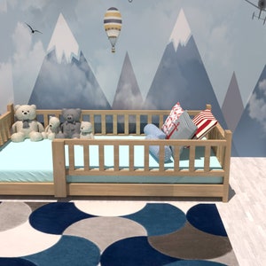 May include: A light wood toddler bed with a blue and white patterned pillow and a blue mattress. The bed is decorated with stuffed animals and is in a room with a white bookshelf and a blue and grey rug.