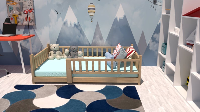 May include: A light wood toddler bed with a blue and white patterned pillow and a blue mattress. The bed is decorated with stuffed animals and is in a room with a white bookshelf and a blue and grey rug.