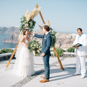 Pu&ograve; includere: Una cerimonia di matrimonio all'aperto con una coppia in piedi sotto un arco di legno decorato con erba della pampa e vegetazione. La cerimonia si svolge su una scogliera con vista sull'oceano.