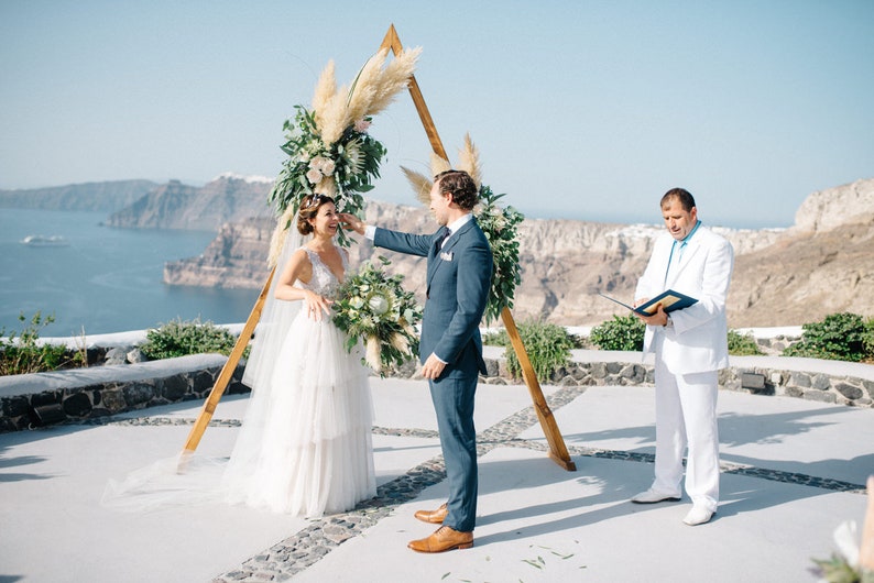 Pu&ograve; includere: Una cerimonia di matrimonio all'aperto con una coppia in piedi sotto un arco di legno decorato con erba della pampa e vegetazione. La cerimonia si svolge su una scogliera con vista sull'oceano.