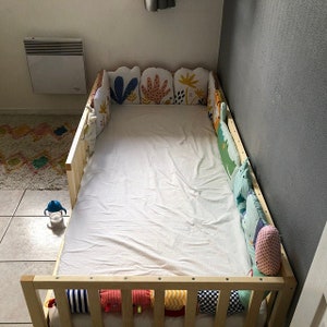 May include: A white toddler bed with a wooden frame and colourful patterned cushions around the perimeter. The cushions are made of fabric with a variety of colours and patterns, including floral, geometric, and animal prints.