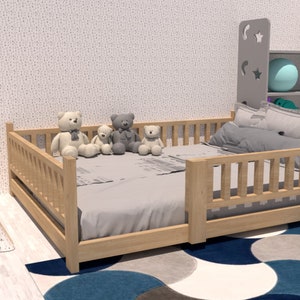 May include: A wooden toddler bed with a grey mattress and a grey and white patterned sheet. Four teddy bears are sitting on the bed. The bed has a wooden railing around it. The bed is in a room with a white wall and a blue and grey rug.