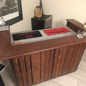 May include: A wooden bar with a dark brown stain. The bar has a built-in shelf and a raised lip on the front. There are two bar towels on the surface, one with the text "JIM BEAM" and the other with the text "BACARDI". There are two clear glasses and a bottle of liquor on the bar.