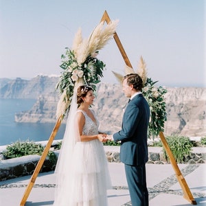Pu&ograve; includere: Una cerimonia di matrimonio all'aperto con una coppia in piedi davanti a un arco triangolare in legno decorato con erba della pampa e vegetazione. La coppia si scambia i voti con una vista panoramica sull'oceano sullo sfondo.