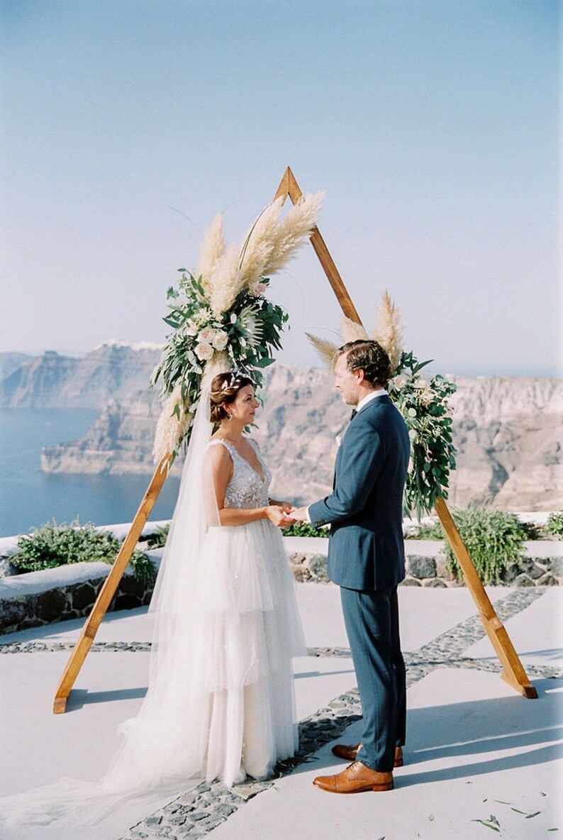 Pu&ograve; includere: Una cerimonia di matrimonio all'aperto con una coppia in piedi davanti a un arco triangolare in legno decorato con erba della pampa e vegetazione. La coppia si scambia i voti con una vista panoramica sull'oceano sullo sfondo.