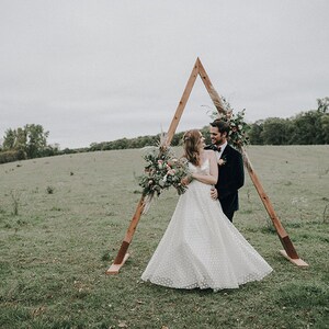 Pu&ograve; includere: Una coppia posa per una foto di matrimonio davanti a un arco triangolare in legno decorato con fiori e piante verdi. La coppia &egrave; in piedi in un campo con erba verde e alberi sullo sfondo.
