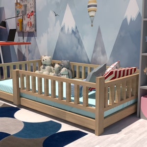 May include: A wooden toddler bed with a blue and white patterned mattress and a white, blue, and red striped pillow. The bed is surrounded by a wooden railing and has a few stuffed animals sitting on it. The bed is in a room with a blue and grey mountain-themed wallpaper.