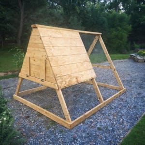 Puede incluir: Un gallinero de madera en forma de A, con techo inclinado y laterales de malla metálica. El gallinero tiene una pequeña puerta y está construido con madera de color claro. Se encuentra sobre una superficie de grava, con árboles al fondo.