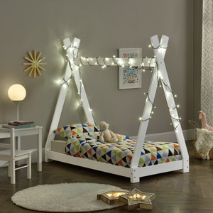 May include: White wooden teepee bed frame with string lights draped across the top. The bed is made with a colorful patterned duvet and a stuffed animal is sitting on the bed.