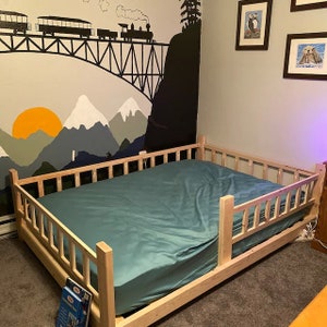 May include: A wooden toddler bed with a teal mattress and a mountain and train mural on the wall behind it.