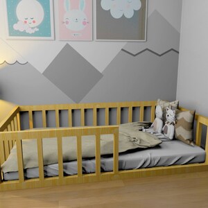 May include: A wooden toddler bed with a safety rail on one side. The bed is made of natural wood and has a grey mattress. There are two stuffed animals on the bed, a grey bunny and a grey cat.