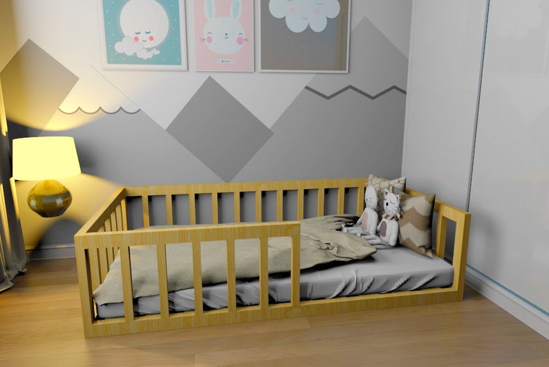May include: A wooden toddler bed with a safety rail on one side. The bed is made of natural wood and has a grey mattress. There are two stuffed animals on the bed, a grey bunny and a grey cat.
