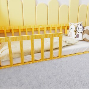 May include: A yellow wooden toddler bed with a white mattress and a white and brown patterned pillow. The bed is on a white fluffy rug.