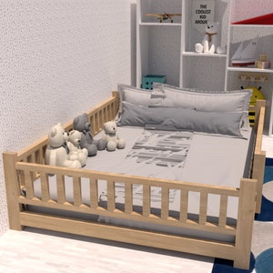 May include: A wooden toddler bed with a grey and white patterned bedding set. Four plush teddy bears are sitting on the bed. The bed has a wooden railing around the perimeter. The bed is in a room with a white wall and a white bookshelf with books and toys.
