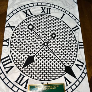 May include: White fabric with a black and white clock design. The clock face has Roman numerals and a black and white pattern. The fabric has the text "Guaranteed Real Dutch Block Prints" and "Printed in Holland".