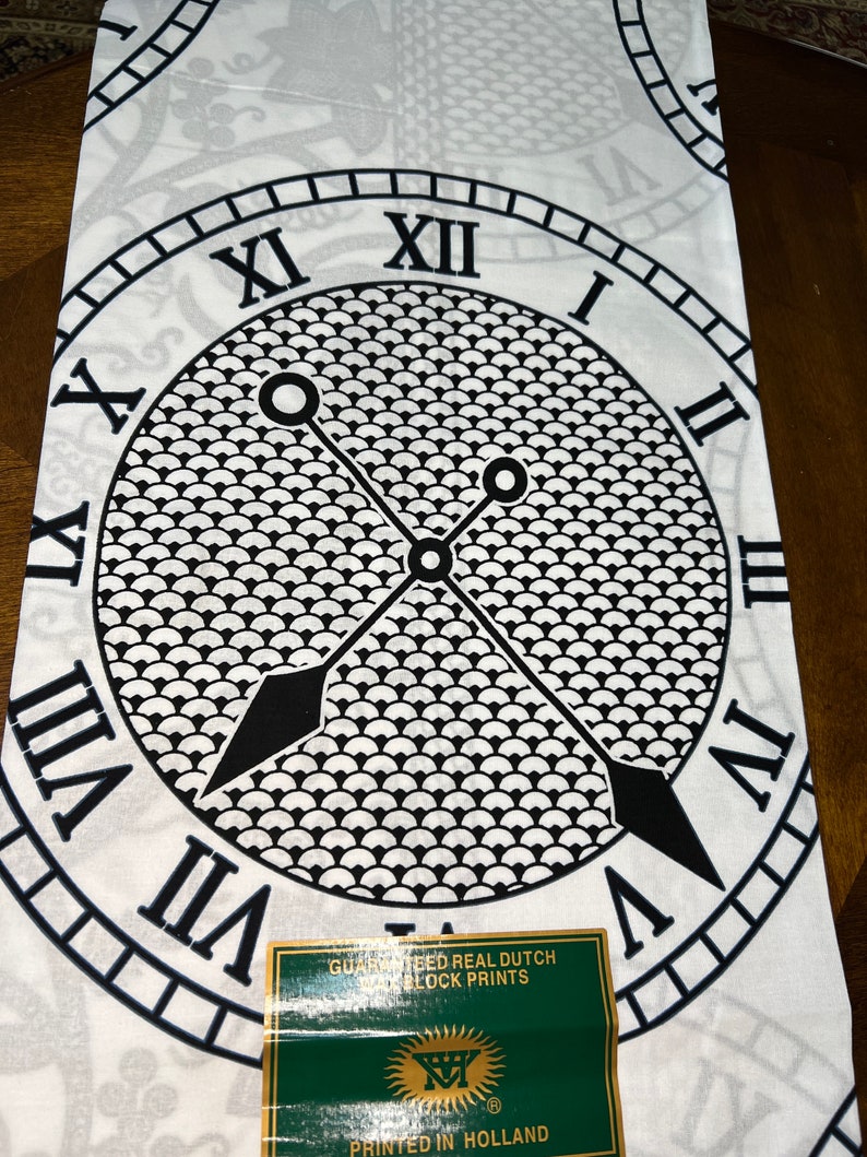 May include: White fabric with a black and white clock design. The clock face has Roman numerals and a black and white pattern. The fabric has the text "Guaranteed Real Dutch Block Prints" and "Printed in Holland".