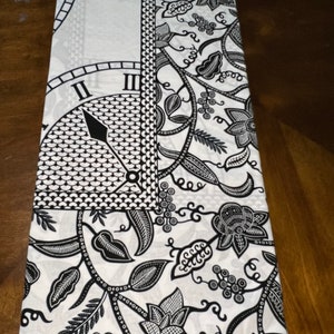 May include: Black and white fabric with a floral pattern and a clock face design. The fabric has a white background with black floral vines, leaves, and flowers. The clock face is in the upper left corner and has black Roman numerals and a black hand.